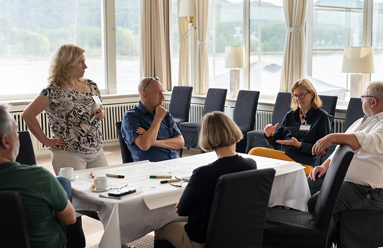 Six participants sit at a table and exchange ideas they have collected. One participant explains an idea in more detail, the rest of the participants listen with attention and interest.